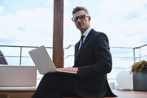 Confident mature man working on laptop while standing on the rooftop terrace photo