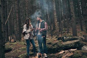 Love inspires them. Full length of beautiful young couple smiling and looking at each other while warming up near the campfire photo