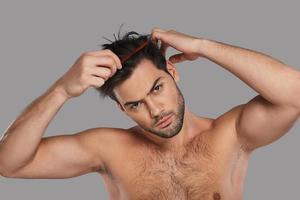 Paying attention to every detail. Good looking young man combing his hair while standing against grey background