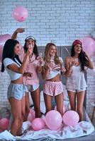 nada es mejor que los amigos. Longitud total de cuatro atractivas jóvenes mujeres sonrientes en pijama bebiendo champán mientras tienen una fiesta de pijamas foto