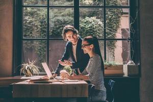Discussing business together. Confident young man and woman looking at laptop and smiling while sitting at the wooden desk in creative office or cafe photo