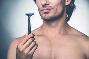 Time for new razor. Close-up young shirtless man holding razor and expressing negativity while standing against grey background photo