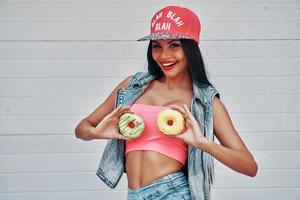 Being young means having fun. Playful young women holding donuts against her breast and smiling while standing against white background photo