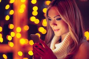 Typing lovely message to him. Beautiful young smiling woman in white sweater holding smart phone and looking at it with defocused Christmas lights in the background photo