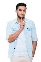 In search of solution. Thoughtful young Indian man touching his chin with finger and looking away while standing against white background photo