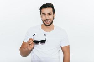 Charming heartbreaker. Handsome young Indian man carrying sunglasses and looking at camera with smile while standing against white background photo