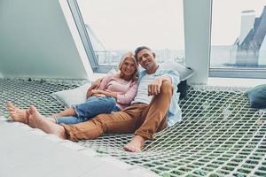 Beautiful mature couple smiling and looking at camera while relaxing in big hammock at home together photo