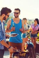 almuerzo de verano perfecto. dos jóvenes alegres haciendo barbacoas y sonriendo mientras dos mujeres hablan entre sí en el fondo foto