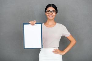 Copy space on her clipboard. Beautiful young businesswoman stretching out clipboard and looking at camera while standing against grey background photo