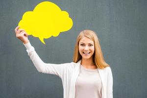 Fresh idea. Beautiful young woman holding empty speech bubble and looking at camera while standing against grey background photo