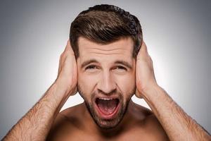 Too loud sound. Portrait of frustrated young shirtless man shouting and covering ears by hands while standing against grey background photo