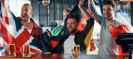 animando a los jóvenes cubiertos de banderas internacionales disfrutando de la cerveza mientras ven un partido deportivo en el pub foto