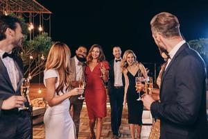 grupo de personas hermosas en ropa formal comunicándose y sonriendo mientras pasan tiempo en una fiesta de lujo foto