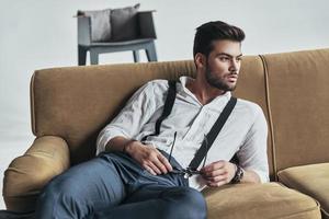 Thinking about something important. Stylishly dressed young man holding his eyewear and looking away while lying on sofa photo