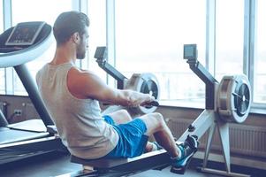 Rowing to success. Side view of young man in sportswear doing rowing in front of window at gym photo