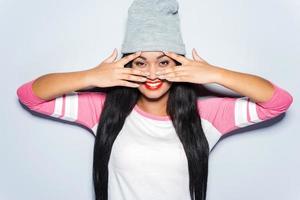 Just fun in my mind. Happy young African woman in funky clothes touching her face with fingers and smiling while standing against grey background photo