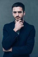 Confident business expert. Thoughtful young man holding hand on chin and looking at camera while standing against grey background photo