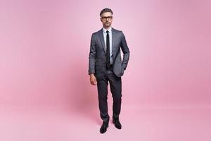 Confident mature man in full suit looking at camera while standing against pink background photo