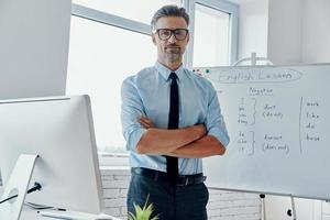 Confident English teacher looking at camera while standing near the whiteboard at classroom photo