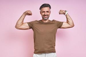 Happy mature man showing his biceps while standing against pink background photo