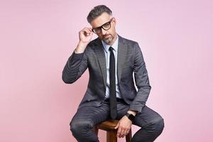 Handsome mature man in formalwear adjusting eyeglasses while sitting against pink background photo