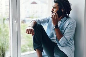 encantador joven africano hablando por teléfono inteligente mientras se sienta en el alféizar de la ventana en el interior foto