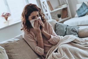 Sneezing non-stop. Top view of sick young women blowing her nose using facial tissues while sitting on the sofa at home photo