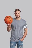 Handsome young man carrying a basketball ball and looking at camera while standing against grey background photo