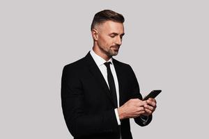 Always available. Good looking young man in full suit using his smart phone and smiling while standing against grey background photo