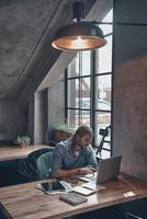 Feeling passionate about his project. Top view of thoughtful young man working using computer while sitting at the office desk near the window photo