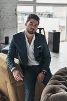 Elegance and masculinity. Thoughtful young man in full suit looking at camera while leaning on the sofa photo