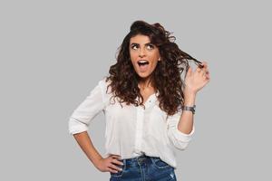 Playful girl. Attractive young woman playing with her hair and keeping mouth open while standing against grey background photo