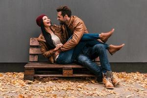 Carefree time together. Beautiful young couple having fun together while sitting on the wooden pallet together with grey wall in the background and fallen leaves on ht floor photo