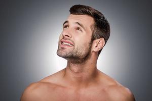 Lost in thoughts. Portrait of thoughtful young shirtless man looking up and smiling while standing against grey background photo