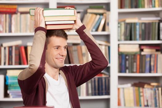 Knowledge in my head. Cheerful young man holding a book stack on his head and smiling while sitting at the library photo