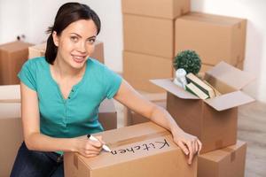 Writing on a carton box. Beautiful young woman marking a cardboard box and smiling at camera while more boxes laying on background photo