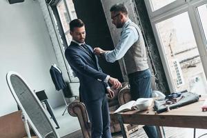 Talented tailor. Young fashionable designer helping his client to get dressed while standing in his workshop photo