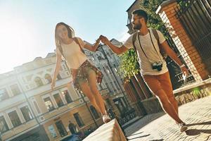 toda la longitud de una pareja joven juguetona tomándose de la mano y sonriendo mientras camina al aire libre foto