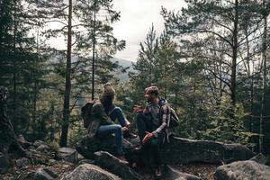 Finally enjoying the travel. Beautiful young couple sitting on the rocks and talking while hiking together in the woods photo