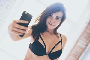 Selfie in the morning. Beautiful young woman in black lingerie making selfie while standing in front of window photo