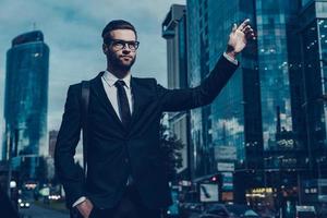 Catching taxi. Night time image of confident young businessman in full suit catching taxi while raising his arm and standing outdoors with cityscape in the background photo