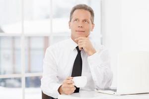 In search of the new ideas. Thoughtful mature man in formalwear sitting at his working place and drinking coffee photo