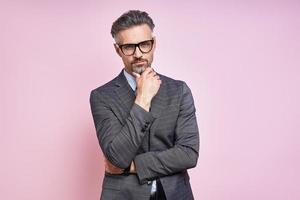 Handsome mature man in formalwear holding hand on chin while standing against pink background photo