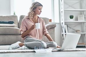 Coffee brings fresh ideas. Beautiful young woman holding a cup and using laptop while sitting on the floor at home photo