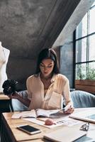 Always aware of latest fashion news. Beautiful young woman looking at fashion magazine and holding digital camera while sitting in her workshop photo