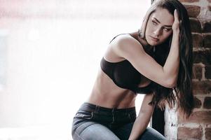 Feeling lonely. Beautiful young thoughtful woman in bra and jeans looking away while sitting at the window sill photo