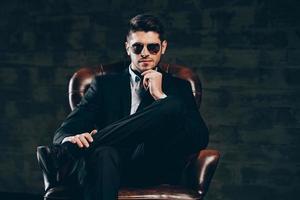 All about style. Young handsome man in suit and sunglasses holding hand on chin and looking at camera while sitting in leather chair against dark grey background photo