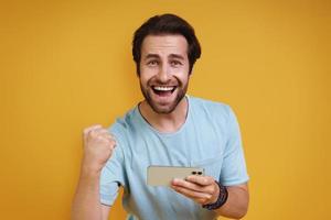 Happy young man in casual clothing holding smart phone and gesturing while standing against yellow background photo