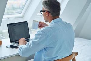 Rear view of confident mature man working on laptop while sitting at the office desk photo
