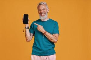 Happy senior man pointing smart phone and smiling while standing against orange background photo